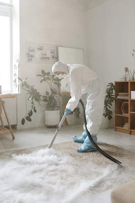 A person in a white protective suit, face mask, and blue gloves is using a professional vacuum cleaner to deep clean a beige, textured carpet in a well-lit, modern living room. The room features white