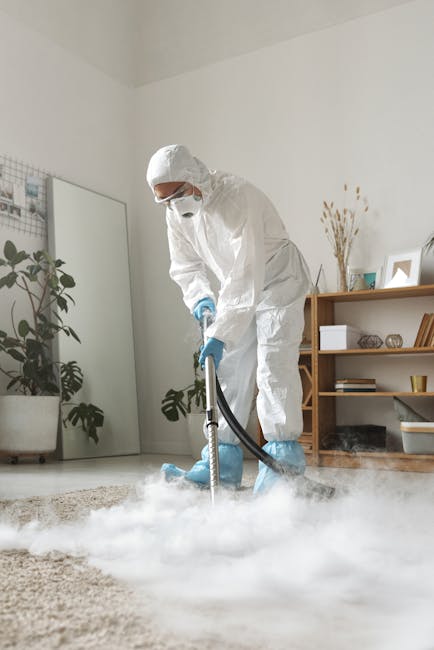 A person in a white protective suit, face mask, and blue gloves is using a professional vacuum cleaner to deep clean a beige, textured carpet in a well-lit, modern living room. The room features white