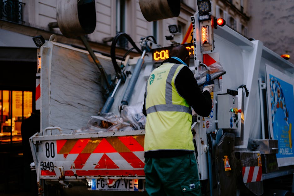 An outdoor scene featuring a street cleaner wearing a bright green uniform, including a cap and jacket, standing next to a large green waste bin. The worker is holding a bundle of dried twigs and broo
