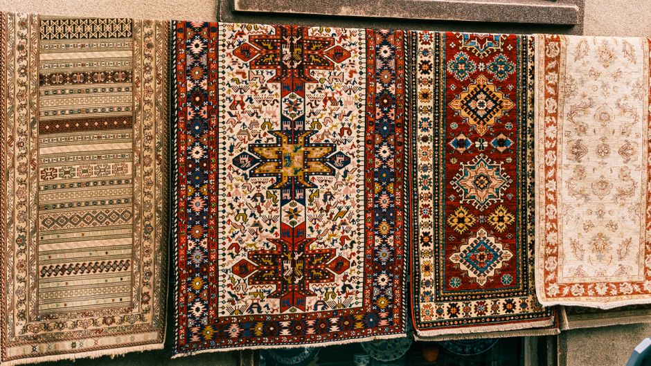 A side-by-side comparison image of two Persian-style area rugs displayed in a shop window, showing a before and after case study of deep cleaning by Hackney Carpet Cleaners. The left rug appears dull,